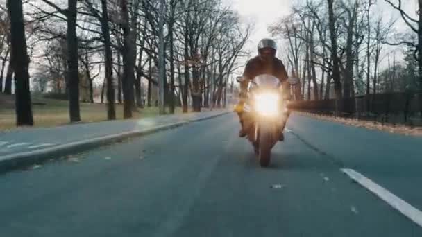 Байкер в шлеме с открытым козырьком едет на мотоцикле к камере вдоль городского парка — стоковое видео
