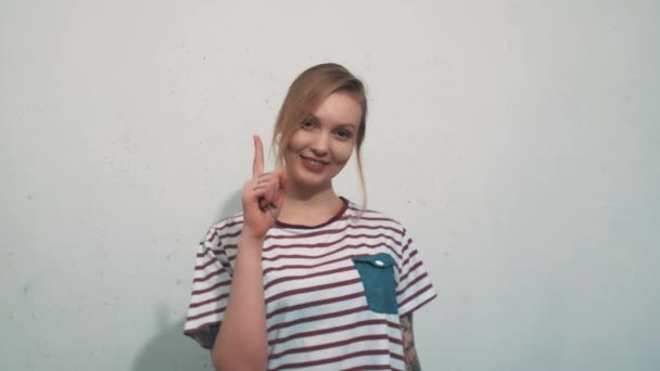 Csinos, fiatal szőke lány húzza fel egy ujjal, fehér fal előtt — Stock videók