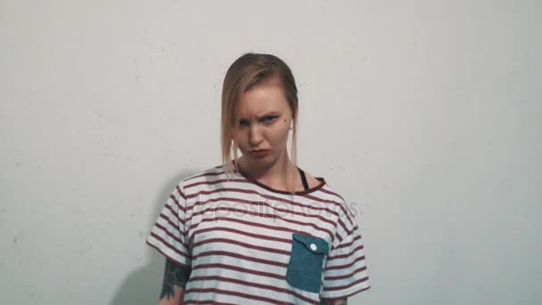 Chica rubia gruñona en camisa a rayas con tatuajes enfurruñados delante de la pared blanca — Vídeo de stock