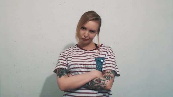 Menina loira irritada em camisa listrada com tatuagens sulcos na frente da parede branca — Vídeo de Stock