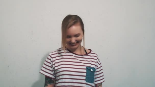 Радостная блондинка в полосатой рубашке с татуировками, смеющаяся перед белой стеной — стоковое видео