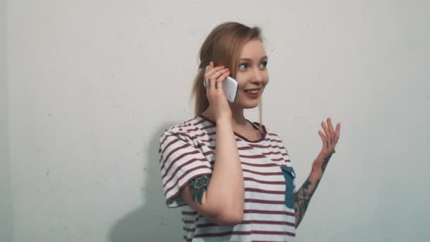 Красива жінка в смугастій сорочці з татуюваннями, що розмовляють на смартфоні на білій стіні — стокове відео