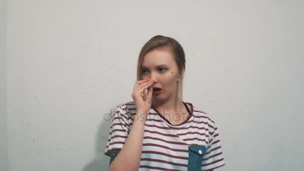 Chica rubia joven tonta en camisa a rayas con tatuajes que rascan la nariz en la pared blanca — Vídeo de stock