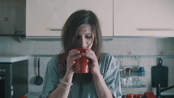 Привлекательная девушка пьет кофе из красной кружки и улыбается на кухне по утрам — стоковое видео