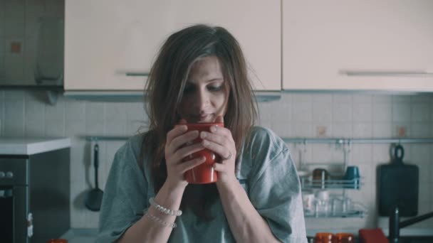 Красивая девушка пьет кофе из красной кружки и улыбается на кухне утром — стоковое видео