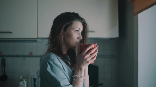 Junges attraktives Mädchen schaut ins Fenster und trinkt morgens Kaffee in der Küche — Stockvideo