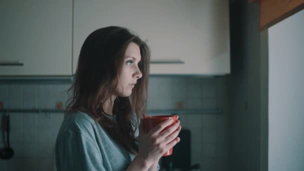 Νεαρή όμορφη γυναίκα ματιά στο παράθυρο και ποτά καφέ στην κουζίνα στο πρωί — Αρχείο Βίντεο