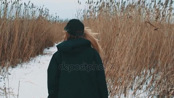 Mujer rubia joven caminando por el camino de la nieve entre cañas secas en el día nublado de invierno — Vídeos de Stock