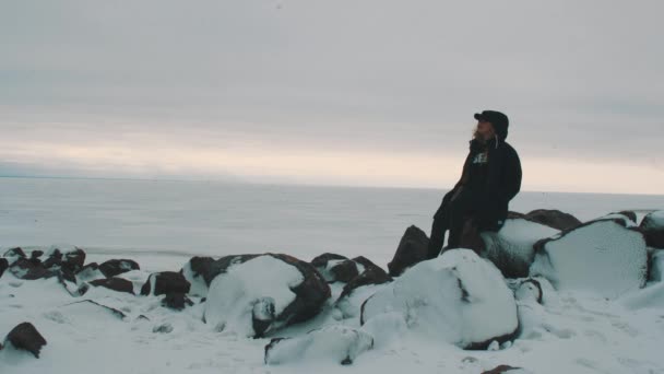 Junge blonde Frau in schwarzer Jacke und Mütze sitzt auf Steinen vor gefrorenem Meer — Stockvideo
