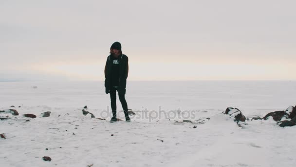 Ung blond kvinne i svart jakke og lue stående på frossen sjø – stockvideo