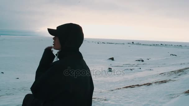 Giovane ragazza silhouette in giacca nera e cappello seduto sulla riva del mare congelato — Video Stock