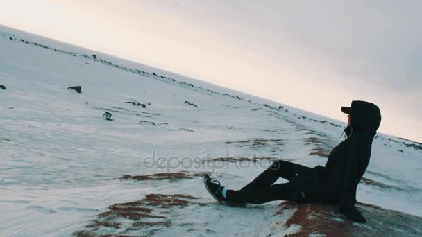 Νεαρή ξανθιά γυναίκα στο μαύρο σακάκι και καπάκι κάθεται στην όχθη της λίμνης παγωμένο — Αρχείο Βίντεο