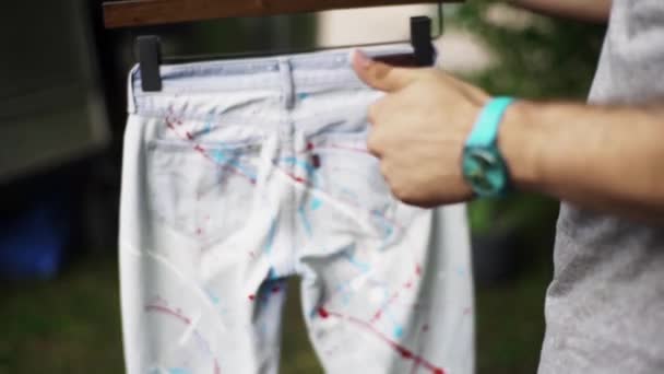 Männliche Hände halten Kleiderbügel mit weißen Jeans, die mit Farbspritzern bedeckt sind — Stockvideo
