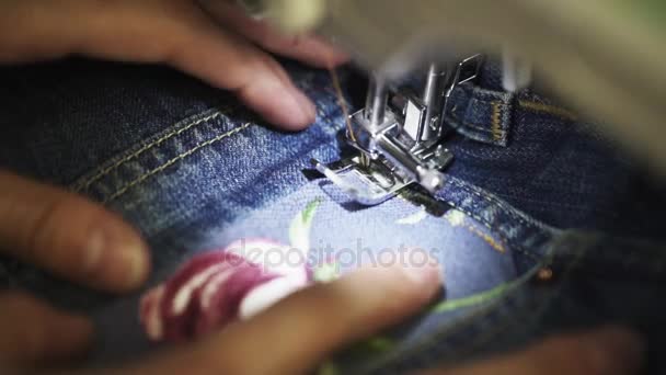 As mãos do alfaiate da mulher usam a máquina de costura para costurar o remendo com imagem da rosa à ganga — Vídeo de Stock