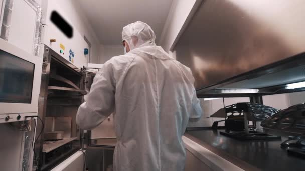 Scientist in robe and rubber gloves puts metal element inside oven in laboratory — Stock Video