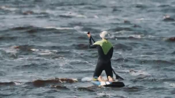 Deportista en traje de baño completo sentado paseos tabla de surf utilizando paleta en un día ventoso — Vídeo de stock