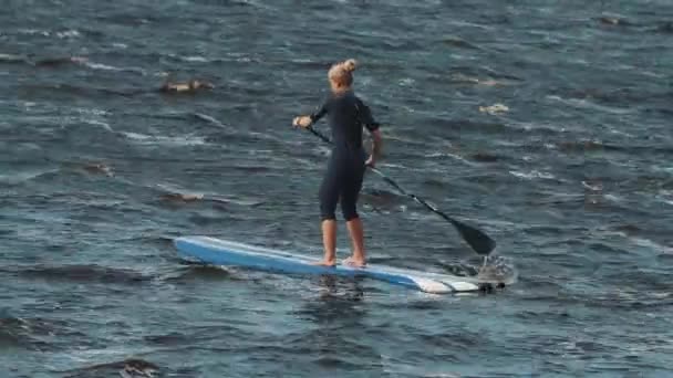Women with hair bun in swimsuit rides a surfboard rowing paddle. Windy day — Stock Video
