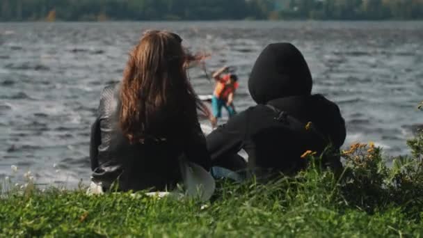 夫妇坐在草地上看着两个冲浪者骑过在他们面前的湖面 — 图库视频影像