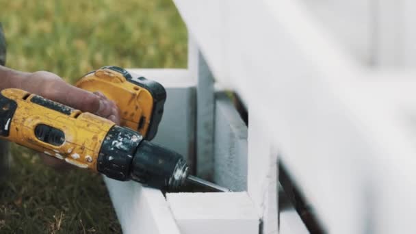 La mano del trabajador utiliza un destornillador eléctrico para ensamblar la estructura de madera sobre la hierba — Vídeos de Stock