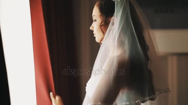 Jonge bruid meisje in trouwjurk opent gordijnen in halve donkere kamer — Stockvideo