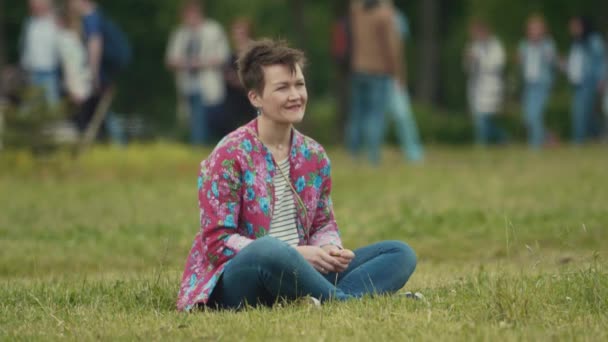 SAINT PETERSBURG, RÚSSIA - JUNHO 24, 2017: Mulher de cabelos curtos em camisa floral sentada na grama no evento de verão do parque — Vídeo de Stock