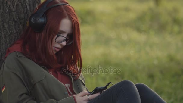 SAINT PETERSBURG, RUSSIE - 24 JUIN 2017 : Jeune rousse écoutant de la musique avec des headhones assise sous un arbre dans un parc — Video
