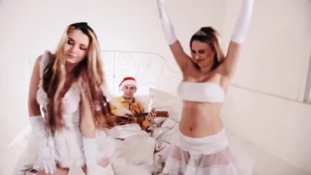 Man in santa hat play ukulele and singing. Girls seductive dancing in big bed — Stock Video