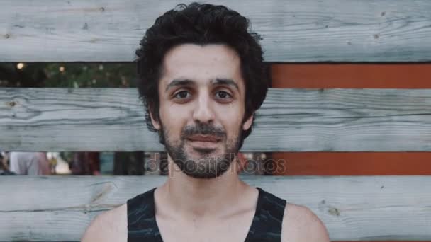 Joven árabe barbudo hombre en tanque superior sonrisas y olas mano a cámara — Vídeos de Stock