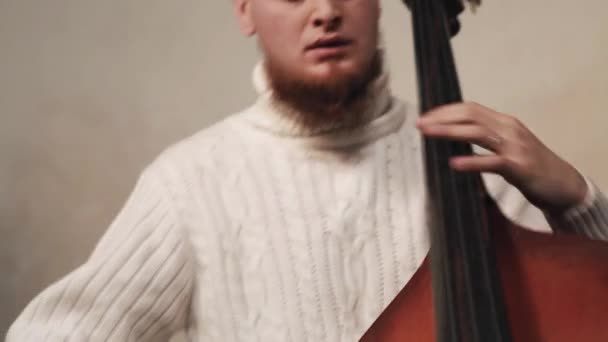 Молодой бородатый мужчина в белом трикотажном свитере играет на контрабасе со скрипачом — стоковое видео