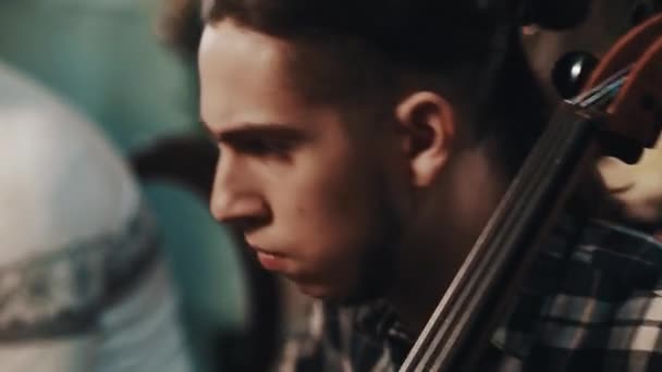Jovem performer com rabo de cavalo em camisa xadrez tocando violoncelo com violinista — Vídeo de Stock