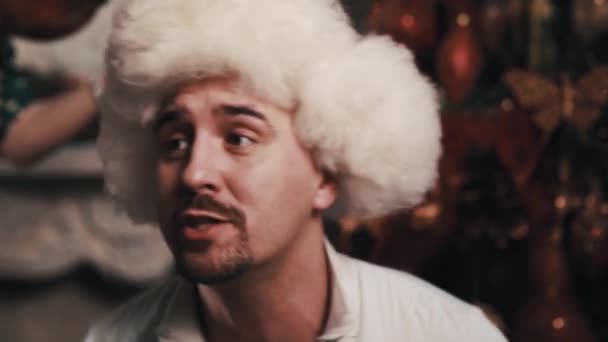 Unshaved vocalist in fur hat perform sitting in room with christmas decorations — Stock Video