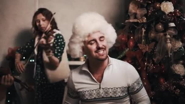 Sångare i päls hatt sång med stråkkvartett som sitter nära julgran — Stockvideo