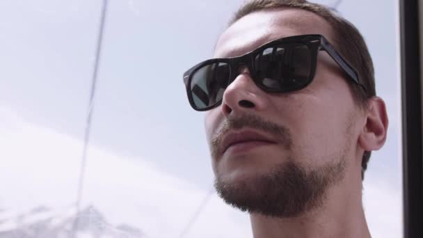 Joven barbudo en gafas de sol montando funitel lift en zona de picos nevados — Vídeos de Stock