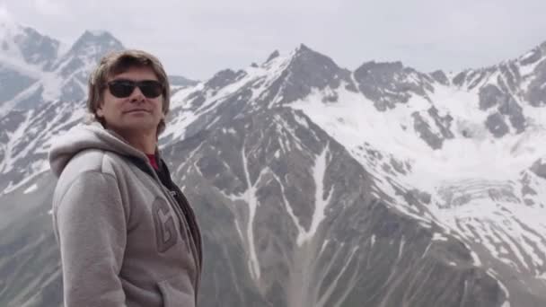 Мужчина средних лет в толстовке и солнцезащитных очках оглядывается вокруг горы — стоковое видео