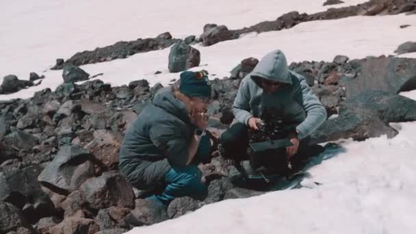 Two men filming crew making camera adjustments at mountains snowy cliff — Stock Video