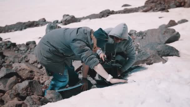 Two men filming crew placing camera on waterproof pad at mountains snowy cliff — Stock Video