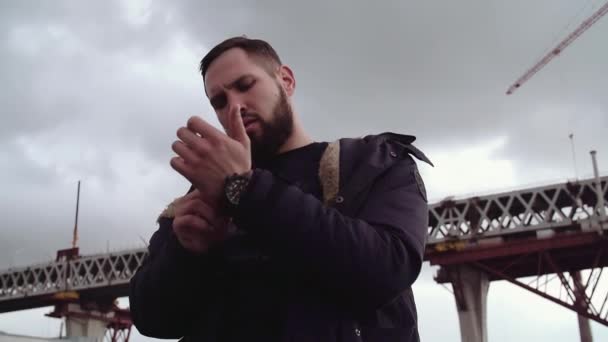 Бородатый молодой человек регулирует наручные часы стоя снаружи под облачным небом — стоковое видео