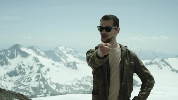 Молодой бородач указывает на камеру в заснеженных горах с живописным видом — стоковое видео