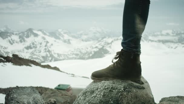 Ноги путешественника в кожаных сапогах топчутся на скале при снежном горном пейзаже — стоковое видео