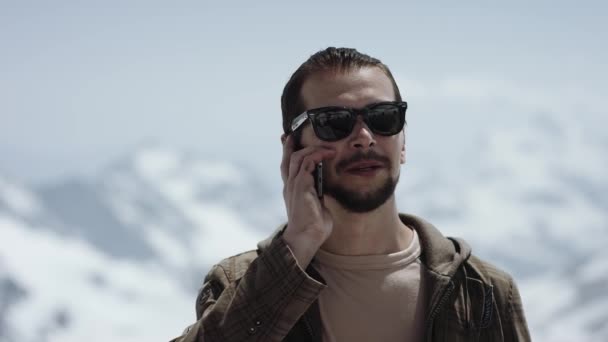 Молодой бородатый человек на вершине горы с живописным видом разговаривает по мобильному телефону — стоковое видео