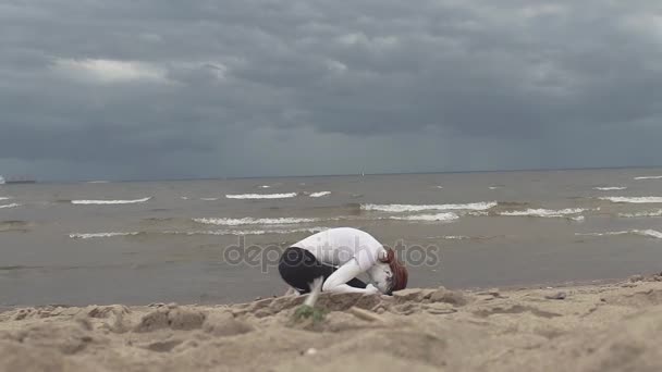 Художественная женщина, покрытая белой краской, присела на песчаном берегу моря — стоковое видео