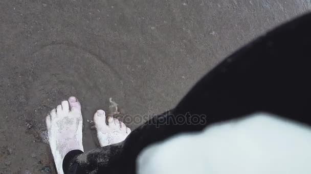 किनारपट्टीवर चालणार्या लाटांमध्ये उभे पांढरा पेंटमध्ये झाकलेल्या कलात्मक स्त्रीचे पाय — स्टॉक व्हिडिओ