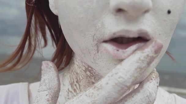 Καλλιτεχνική κορίτσι που καλύπτονται σε λευκό χρώμα σύρετε το δάχτυλο πάνω από τα χείλη στην ακτή της θάλασσας — Αρχείο Βίντεο