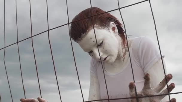 Performance girl in white paint leans on cage on sea shore along metal fence — Stock Video