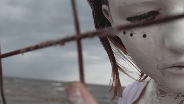 Rendimiento femenino en pintura blanca se apoya en la jaula en la orilla del mar a lo largo de valla metálica — Vídeo de stock