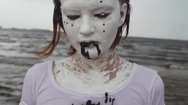 Performance artist woman covered in white paint drools black liquid on sea shore — Stock Video