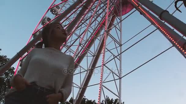 Pretty young girl in glasses standing under ferris wheel with flashing lights — Stock Video