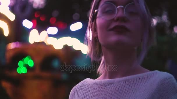 Красивая девушка в очках позирует в парке аттракционов рядом с аттракционом — стоковое видео