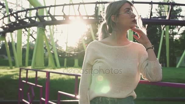 Красивая молодая девушка в очках, стоящая возле американских горок в яркой солнечной вспышке — стоковое видео