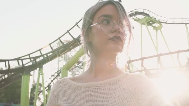 Красивая молодая девушка в очках, стоящая возле американских горок в ярком солнечном свете — стоковое видео